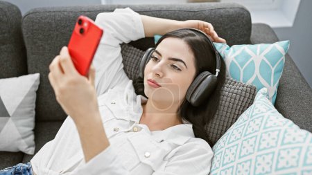 A beautiful young woman enjoys music on headphones while browsing her smartphone in a cozy living room.