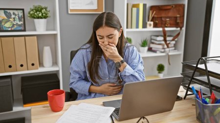 Foto de Mujer hispana joven sintiendo dolor de estómago mientras trabaja en su escritorio de la oficina, expresando malestar. - Imagen libre de derechos