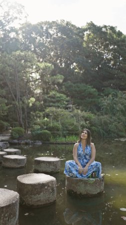 Schöne hispanische Frau sitzt auf einem steinernen Pfad und genießt die grüne Sommerpracht der Natur an einem ruhigen See bei Heian Jingu, Kyoto - eine göttliche Reise, bei der man sich im traditionellen japanischen Tempel umsieht