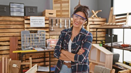 Photo for Smiling woman with safety goggles stands confidently in a cluttered carpentry workshop, embodying skilled labor. - Royalty Free Image
