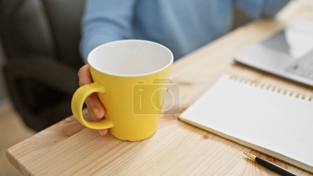 Téléchargez les photos : Gros plan de la main d'une femme tenant une tasse jaune sur un bureau en bois avec un ordinateur portable et un bloc-notes en arrière-plan suggérant une pause du travail. - en image libre de droit