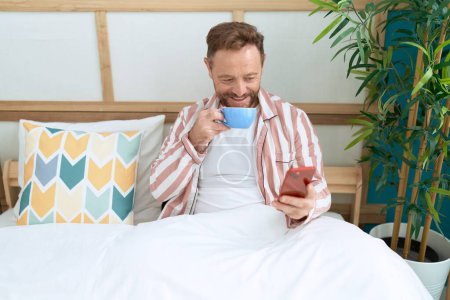 Téléchargez les photos : Homme du moyen âge en utilisant smartphone boire du café dans la chambre - en image libre de droit