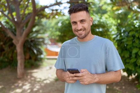 Foto de Joven hombre hispano sonriendo confiado usando smartphone en el parque - Imagen libre de derechos
