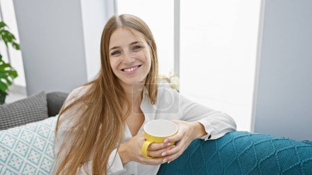 Téléchargez les photos : Une femme souriante aux cheveux blonds s'assoit confortablement à l'intérieur, tenant une tasse de café dans un cadre confortable à la maison. - en image libre de droit