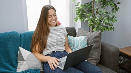 Téléchargez les photos : Une jeune femme souriante aux cheveux bruns utilisant un ordinateur portable sur un canapé sarcelle à l'intérieur. - en image libre de droit
