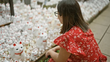 Stunning hispanic woman, glasses adorned, basking in the charm of gotokuji temple's beckoning lucky cat statue in tokyo
