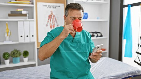 Téléchargez les photos : Homme mûr en blouse buvant du café et vérifiant smartphone dans la chambre du personnel d'une clinique. - en image libre de droit