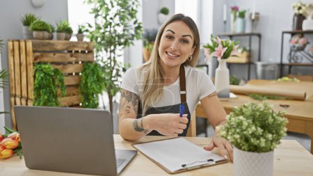 Photo for A smiling woman with tattoos working on a clipboard in a bright flower shop with a laptop and fresh flowers in the background. - Royalty Free Image