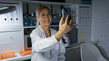 Foto de Una mujer sonriente con una bata de laboratorio se toma una selfie en un entorno moderno de laboratorio. - Imagen libre de derechos