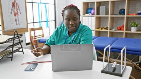 Foto de Mujer afroamericana con trenzas tomando notas en un portátil en una clínica de fisioterapia sala interior - Imagen libre de derechos