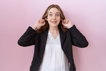 Foto de Joven mujer de negocios caucásica vistiendo chaqueta negra sonriendo tirando de las orejas con los dedos, gesto divertido. problema de audición - Imagen libre de derechos