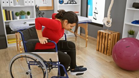 Téléchargez les photos : Une jeune femme hispanique en fauteuil roulant a l'air affligée dans une clinique de réadaptation, avec un équipement thérapeutique visible. - en image libre de droit