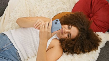 Foto de Una joven hispana con el pelo rizado cómodamente usando un smartphone en una acogedora sala de estar. - Imagen libre de derechos