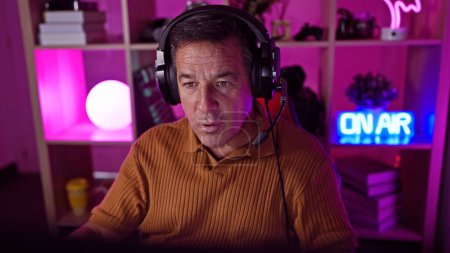 Photo for A middle-aged man wearing headphones focuses intently in a vibrant home gaming room with neon lights at night. - Royalty Free Image