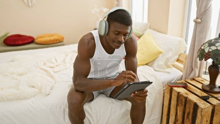 Photo for Smiling african man wearing headphones using tablet in a cozy bedroom setting - Royalty Free Image