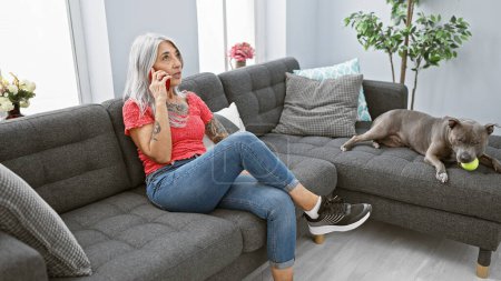 Frau mittleren Alters mit grauen Haaren, tief im Gespräch am Telefon, sitzt gemütlich mit ihrem Hund auf dem gemütlichen Sofa.
