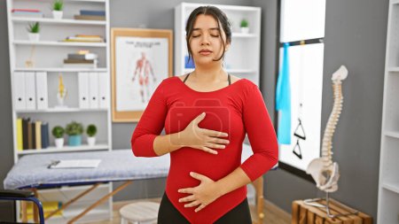 Photo for Young hispanic woman practices deep breathing techniques in a clinic's rehabilitation room, exhibiting a sense of calm and focus - Royalty Free Image