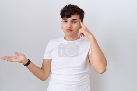 Téléchargez les photos : Jeune homme non binaire portant un t-shirt blanc décontracté confus et agacé avec la paume ouverte montrant l'espace de copie et pointant du doigt vers le front. Penses-y.. - en image libre de droit