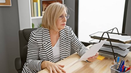 Foto de Una mujer profesional de mediana edad con cabello rubio revisa documentos en un entorno de oficina moderno. - Imagen libre de derechos