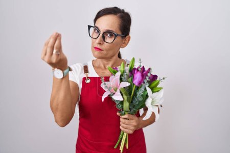 Foto de Mujer morena de mediana edad usando delantal trabajando en floristería sosteniendo ramo haciendo gesto italiano con la mano y los dedos expresión confiada - Imagen libre de derechos
