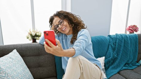 Téléchargez les photos : Femme hispanique d'âge moyen souriante aux cheveux bouclés profitant de la technologie tout en prenant un selfie à l'intérieur sur un canapé confortable. - en image libre de droit