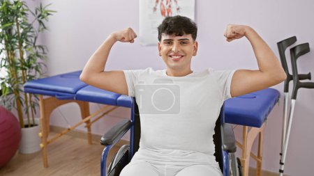 Photo for A smiling young man in a wheelchair flexing muscles confidently in a rehab clinic's bright room. - Royalty Free Image