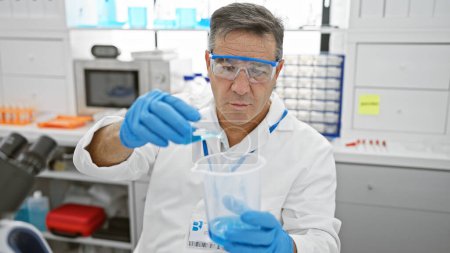 Foto de Un hombre de mediana edad con gafas de seguridad y bata de laboratorio lleva a cabo un experimento en un entorno de laboratorio clínico. - Imagen libre de derechos