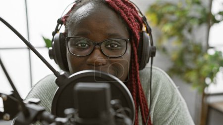 Afroamerikanerin mit Zöpfen trägt Kopfhörer in einem Radiostudio, das für eine Sendung eingerichtet ist.
