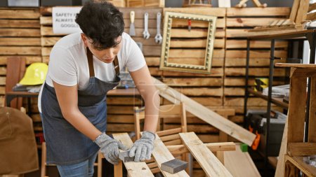 Photo for A young adult man is sanding wood in a well-organized carpentry workshop, showcasing craftsmanship and manual labor. - Royalty Free Image