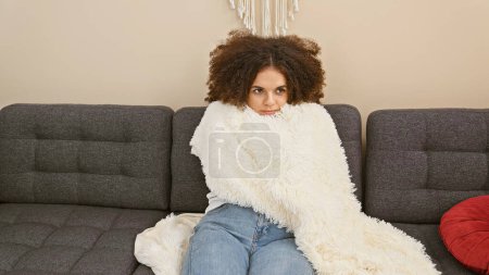Téléchargez les photos : Jeune femme hispanique aux cheveux bouclés enveloppée dans une couverture blanche assise sur un canapé gris à l'intérieur. - en image libre de droit
