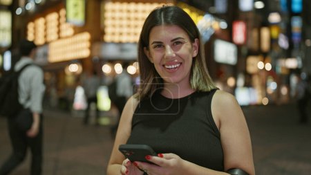 Beautiful hispanic woman smiling at tokyo's urban night lights while texting happily on her smartphone