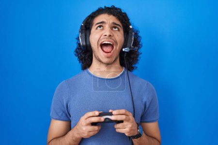 Téléchargez les photos : Homme hispanique avec les cheveux bouclés jouant le contrôleur de tenue de jeu vidéo en colère et fou hurlant frustré et furieux, criant avec colère levant les yeux. - en image libre de droit