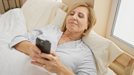Téléchargez les photos : Une femme sereine d'âge moyen se détend au lit avec un smartphone dans une chambre confortable, incarnant loisirs et connectivité moderne. - en image libre de droit