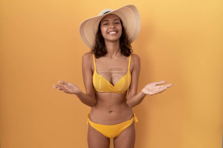 Téléchargez les photos : Jeune femme hispanique portant un bikini et un chapeau d'été souriant et joyeux offrant des mains donnant assistance et acceptation. - en image libre de droit