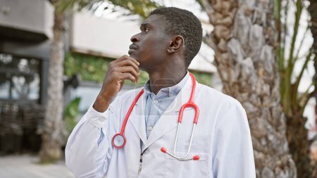 Téléchargez les photos : Un Africain contemplatif vêtu d'un manteau de médecin avec stéthoscope se tient à l'extérieur dans un cadre urbain. - en image libre de droit