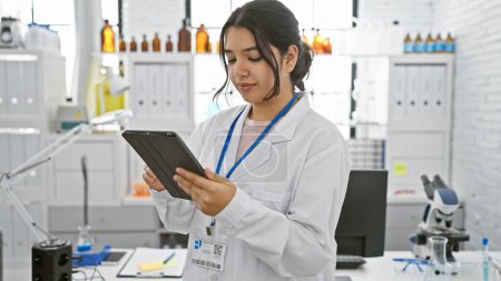 Foto de Mujer hispana en bata de laboratorio examina tableta en ambiente de laboratorio moderno. - Imagen libre de derechos