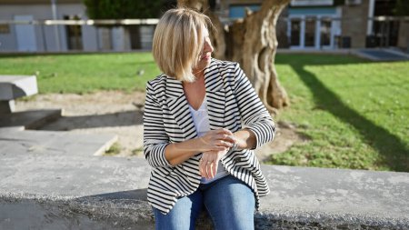 A middle-aged blonde woman sits pensively in a park, exuding a casual elegance against a green outdoor backdrop.