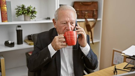 Téléchargez les photos : Au cœur d'un bureau, un homme d'affaires sénior, détendu mais sérieux, parle avec éloquence sur son smartphone, sirotant du café aromatique. - en image libre de droit