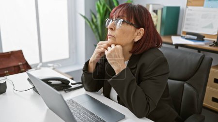 Téléchargez les photos : Pensif hispanique femme dans le bureau contemplant au bureau avec ordinateur portable, lunettes, casque, classeur, intérieur - en image libre de droit