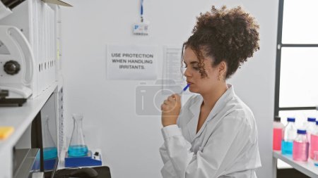 Foto de Mujer hispana pensativa con el pelo rizado enfocado en un entorno de laboratorio, rodeada de equipo científico. - Imagen libre de derechos