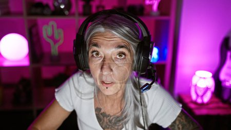 Photo for Mature, grey-haired woman gamer, in serious gaming mode, streaming live in the heart of the night from her cozy home gaming room, sporting headphones and a mic - Royalty Free Image
