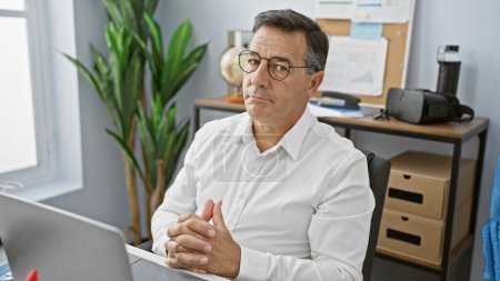 Téléchargez les photos : Un homme contemplatif d'âge moyen portant des lunettes et une chemise blanche, assis dans un cadre de bureau moderne - en image libre de droit
