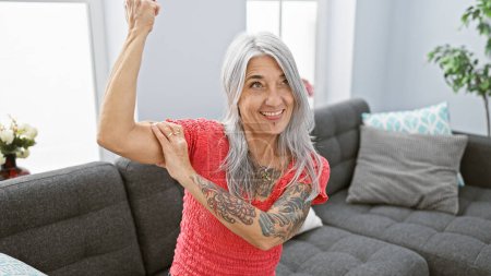 Foto de Mujer de pelo gris, de mediana edad con confianza parpadea gesto fuerte brazo, irradiando alegría y positividad en su acogedora sala de estar en casa. - Imagen libre de derechos