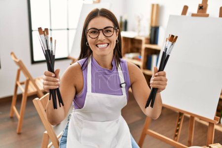 Photo for Young beautiful hispanic woman artist smiling confident holding paintbrushes at art studio - Royalty Free Image