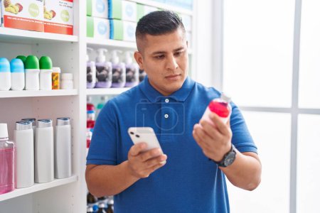 Photo for Young latin man customer using smartphone holding medicine bottle at pharmacy - Royalty Free Image
