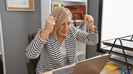 Téléchargez les photos : Femme blonde excitée célébrant le succès au poste de travail de bureau avec ordinateur portable et bloc-notes. - en image libre de droit