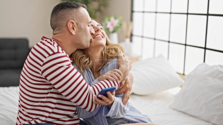 Téléchargez les photos : Homme et femme couple assis sur le lit en utilisant smartphone baisers dans la chambre - en image libre de droit
