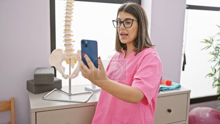 Téléchargez les photos : Une jeune femme hispanique en blouse rose utilise un smartphone dans la salle de désintox d'une clinique. - en image libre de droit