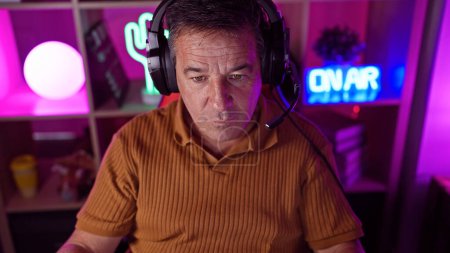 Concentrated middle-aged man wearing headphones in a colorful gaming room at night