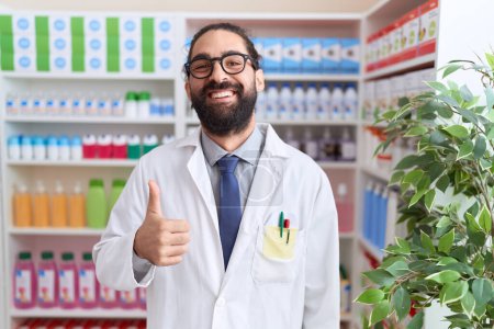 Foto de Hombre hispano con barba trabajando en farmacia sonriendo feliz y positivo, pulgar hacia arriba haciendo excelente y signo de aprobación - Imagen libre de derechos